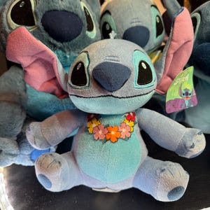 May include: A light blue plush toy character with large dark eyes, prominent ears with pink inner lining, and a colorful floral lei around its neck. The character has a light blue belly patch and a small tag attached to its ear featuring the name Stitch.