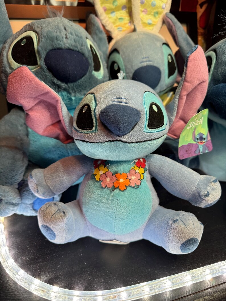 May include: A light blue plush toy character with large dark eyes, prominent ears with pink inner lining, and a colorful floral lei around its neck. The character has a light blue belly patch and a small tag attached to its ear featuring the name Stitch.