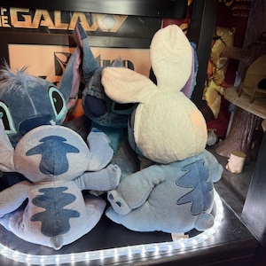 May include: Three blue plush toys resembling the character Stitch. One toy faces forward with large ears, while two others are positioned with their backs to the viewer. One of the rear-facing toys wears a white plush hood with long rabbit ears.