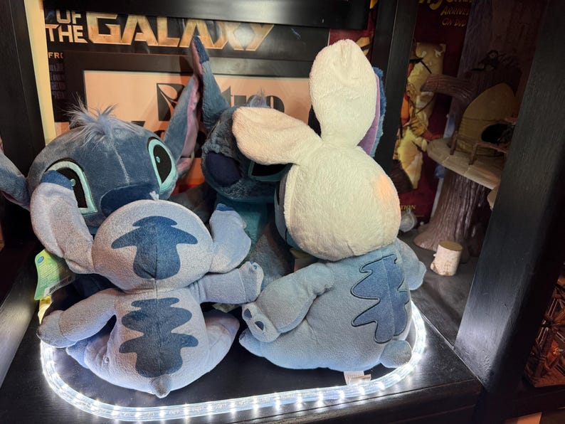 May include: Three blue plush toys resembling the character Stitch. One toy faces forward with large ears, while two others are positioned with their backs to the viewer. One of the rear-facing toys wears a white plush hood with long rabbit ears.