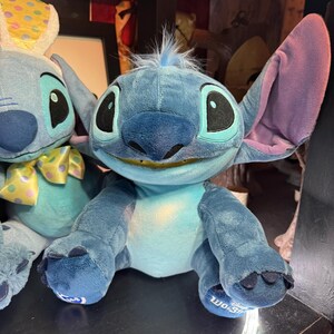 May include: A blue plush toy character with large ears, a wide smile showing teeth, and a tuft of blue hair on its head. The character has dark blue patches on its back and feet, with a lighter blue chest and face. A Build-A-Bear logo is on the foot.