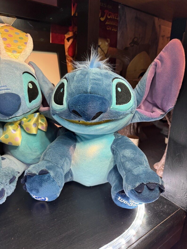 May include: A blue plush toy character with large ears, a wide smile showing teeth, and a tuft of blue hair on its head. The character has dark blue patches on its back and feet, with a lighter blue chest and face. A Build-A-Bear logo is on the foot.