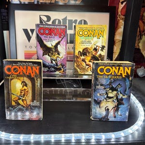 May include: Four vintage paperback books featuring the Conan the Barbarian series. The books have colorful covers with illustrations of Conan in action. Titles include "The Bold," "The Great," "The Unconquered," and "The Victorious."