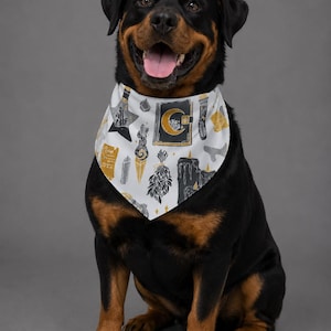 May include: A white dog bandana featuring a repeating pattern of black and gold illustrations including a crescent moon on a book, crystals, candles, and bundles of herbs. The bandana is worn around the neck of a black and tan Rottweiler dog.