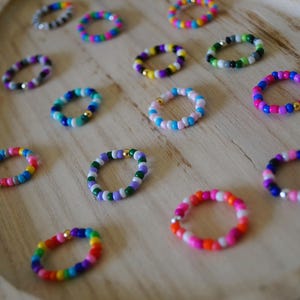 May include: A collection of colorful beaded rings in various sizes and designs. The rings feature a variety of colors, including pink, blue, green, purple, and yellow. Some rings have gold or silver accents. The rings are displayed on a wooden tray.