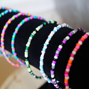 Könnte beinhalten: Eine Sammlung farbenfroher Perlenarmbänder, die auf einem schwarzen zylindrischen Ständer präsentiert werden. Die Armbänder weisen eine Vielzahl von Farben auf, darunter Rosa, Blau, Grün und Weiß, was eine lebendige Darstellung ergibt. Die Perlen sind klein und gleichmäßig verteilt.