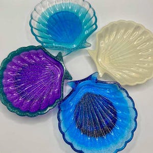 May include: Four seashell-shaped trinket dishes in shades of blue, purple, and cream. The dishes have a glittery finish and are arranged on a white surface. The dishes are ideal for holding jewelry or small items.