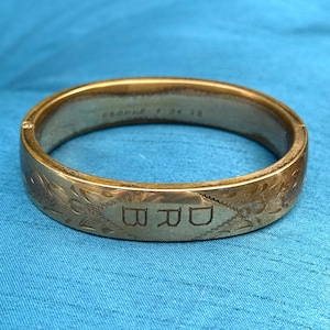 May include: A gold-toned hinged bangle bracelet with engraved floral patterns and the initials "BRD". The bracelet also features the inscription "GEORGE 1 24 73". The jewelry is set against a blue background.
