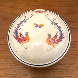 May include: A round, white ceramic trinket box with a lid. The lid features a blue border and is decorated with two colorful birds, a butterfly, and stylized floral elements. The birds have orange, red, and purple plumage.