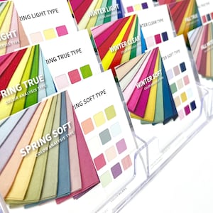 Può includere: Un'esposizione di schede di analisi del colore, ciascuna con uno spettro di tonalità e una palette di colori corrispondente. Le schede sono etichettate con tipi di colore stagionali come "Winter True" e "Spring Soft", e sono disposte in un supporto di plastica trasparente.