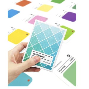 May include: A hand holding a card with a blue and green diamond pattern and the text "COLOR CLASH". Other cards with various colours are in the background, including yellow, pink, and purple. The cards appear to be colour swatches.