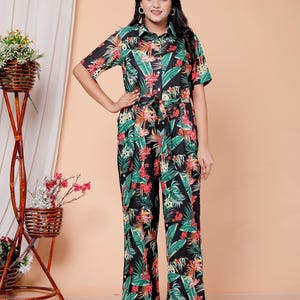Puede incluir: Mono negro con estampado floral tropical con hojas verdes y flores rojas. El mono tiene mangas cortas, cuello camisero y cintura anudada. La modelo lleva sandalias beige.