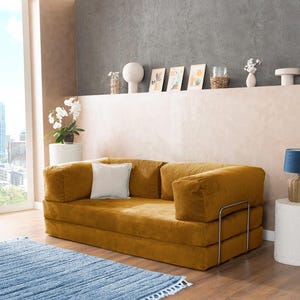 May include: A mustard-colored corduroy sofa with a white pillow, set against a neutral-toned wall. A blue rug with fringe sits on the wooden floor. Decorative items are on a shelf above the sofa.