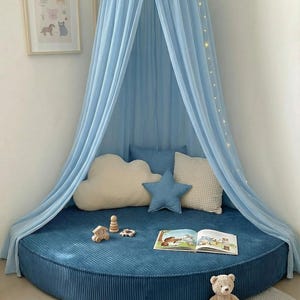 May include: A child's reading nook with a blue canopy and a round, corduroy base. Decorative pillows in cloud and star shapes, a teddy bear, and wooden toys create a playful atmosphere. A children's book is open on the base.