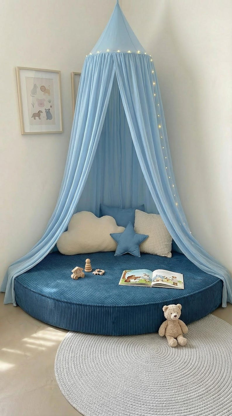 May include: A child's reading nook with a blue canopy and a round, corduroy base. Decorative pillows in cloud and star shapes, a teddy bear, and wooden toys create a playful atmosphere. A children's book is open on the base.