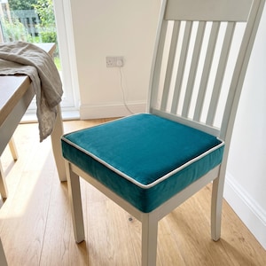 Puede incluir: Una silla de madera blanca con un cojín de asiento cuadrado tapizado en tela de terciopelo color verde azulado. El cojín presenta un ribete blanco en los bordes. La silla tiene un respaldo de listones y cuatro patas rectas de diseño sencillo.