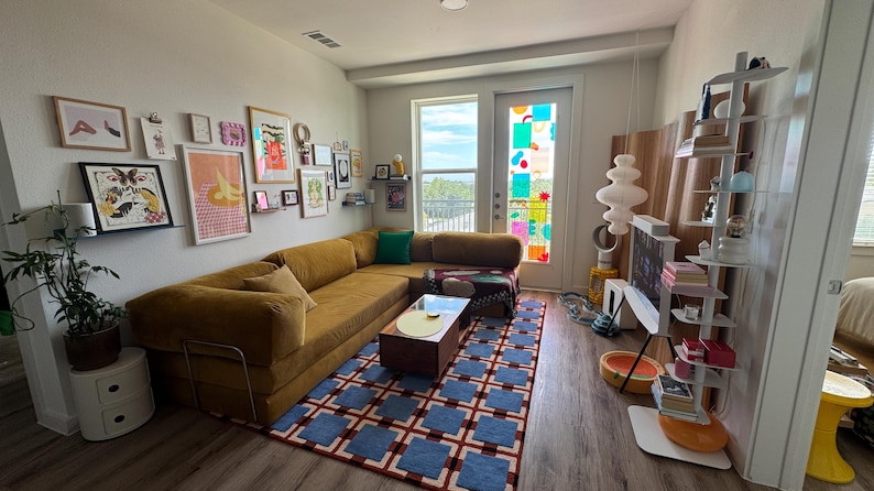 Op de afbeelding: Een moderne woonkamer met een mosterdkleurige hoekbank, een patroonkleed en een verzameling ingelijste kunst aan de muur. Een salontafel staat voor de bank en een decoratieve lamp is zichtbaar.