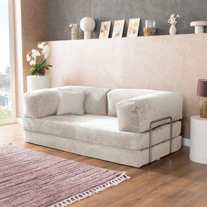 May include: A light beige corduroy sofa with a matching square pillow. The sofa has a low profile and metal arm supports. A mauve rug with a fringed edge is in front of the sofa. The room has a neutral color palette.