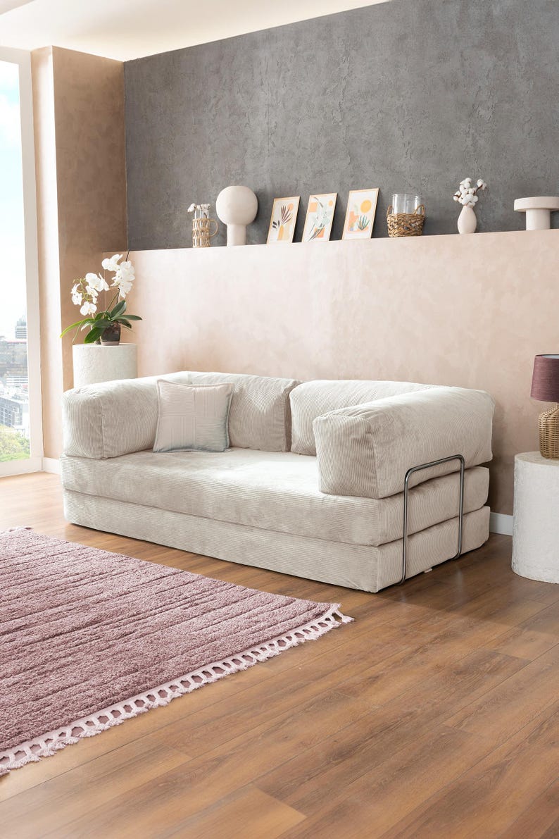 May include: A light beige corduroy sofa with a matching square pillow. The sofa has a low profile and metal arm supports. A mauve rug with a fringed edge is in front of the sofa. The room has a neutral color palette.