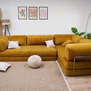 Op de afbeelding: Een grote, mosterdgele hoekbank met verschillende witte kussens. Een ronde, getextureerde, gebroken witte decoratieve bal ligt op de vloer voor de bank. Een houten boekenplank en ingelijste kunstwerken zijn zichtbaar op de achtergrond.