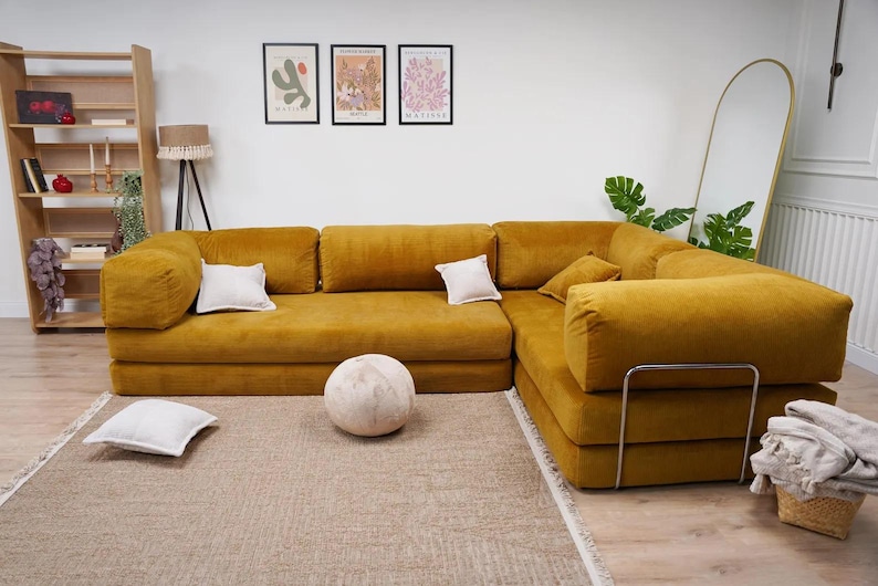 Op de afbeelding: Een grote, mosterdgele hoekbank met verschillende witte kussens. Een ronde, getextureerde, gebroken witte decoratieve bal ligt op de vloer voor de bank. Een houten boekenplank en ingelijste kunstwerken zijn zichtbaar op de achtergrond.