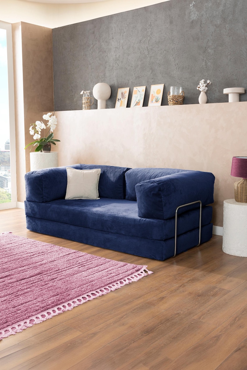 May include: A navy blue corduroy sofa with a white square pillow. A pink rug with a fringed edge sits on a wooden floor. Decorative items are on a shelf in the background.