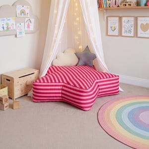 May include: A star-shaped floor cushion featuring horizontal stripes in alternating shades of bright pink and dark red. The cushion is positioned beneath a white fabric canopy with string lights, accompanied by a small cloud and star pillow.