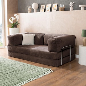 May include: A brown corduroy sofa with a white square pillow. The sofa has a metal frame and is placed on a wooden floor next to a green rug. Decorative items are on a shelf in the background.