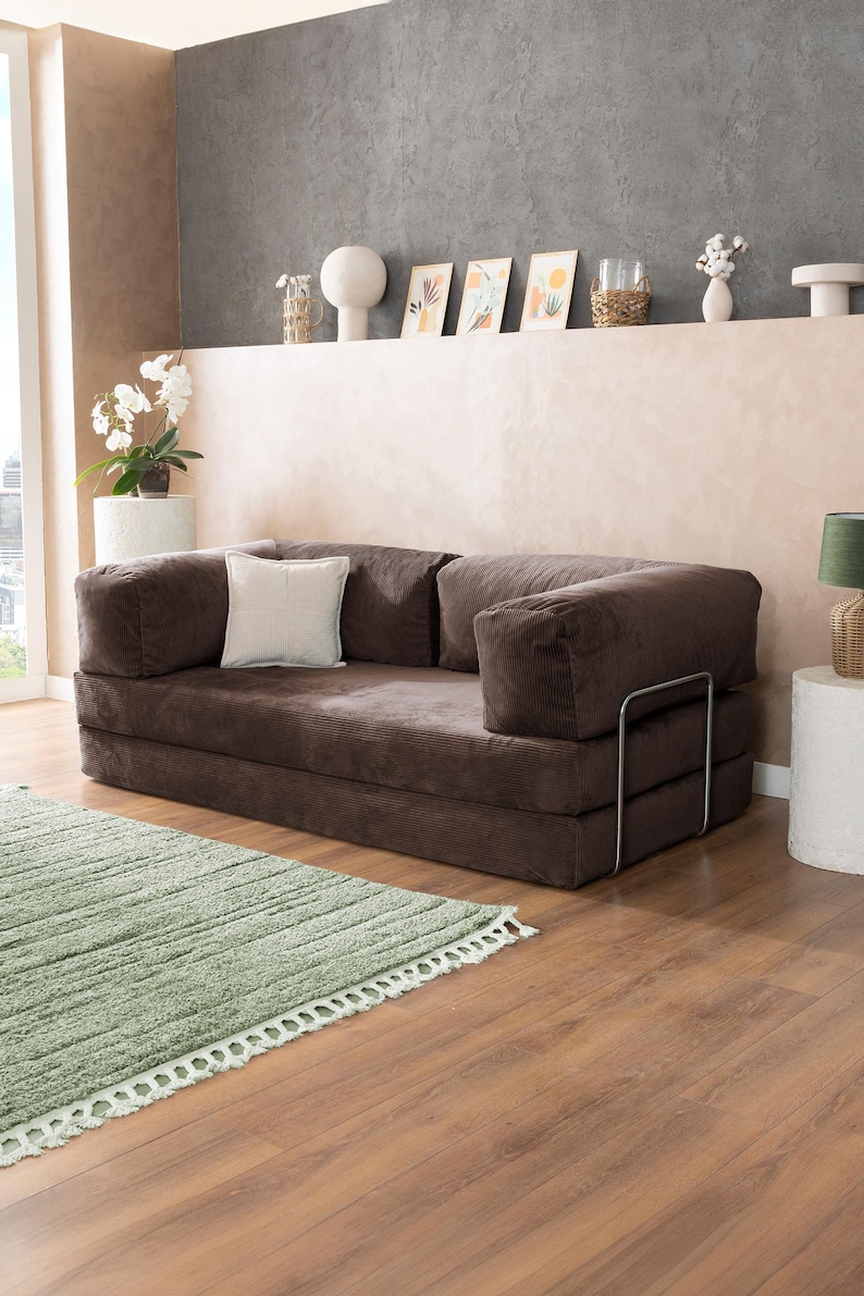 May include: A brown corduroy sofa with a white square pillow. The sofa has a metal frame and is placed on a wooden floor next to a green rug. Decorative items are on a shelf in the background.