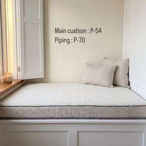 Pode incluir: Uma almofada retangular para banco de janela com tecido texturizado em branco sujo e debrum cinzento, acompanhada por duas almofadas decorativas a condizer. O texto "Main cushion : P-54" e "Piping : P-70" est&aacute; impresso na parede.