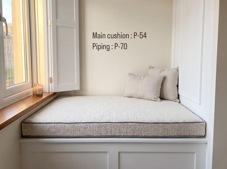 Pode incluir: Uma almofada retangular para banco de janela com tecido texturizado em branco sujo e debrum cinzento, acompanhada por duas almofadas decorativas a condizer. O texto "Main cushion : P-54" e "Piping : P-70" est&aacute; impresso na parede.