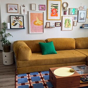 Op de afbeelding: Een mosterdgeel corduroy hoekbank met een groen kussen, een houten salontafel en een blauw en rood patroon tapijt. Verschillende ingelijste kunstwerken hangen aan de witte muur boven de bank.