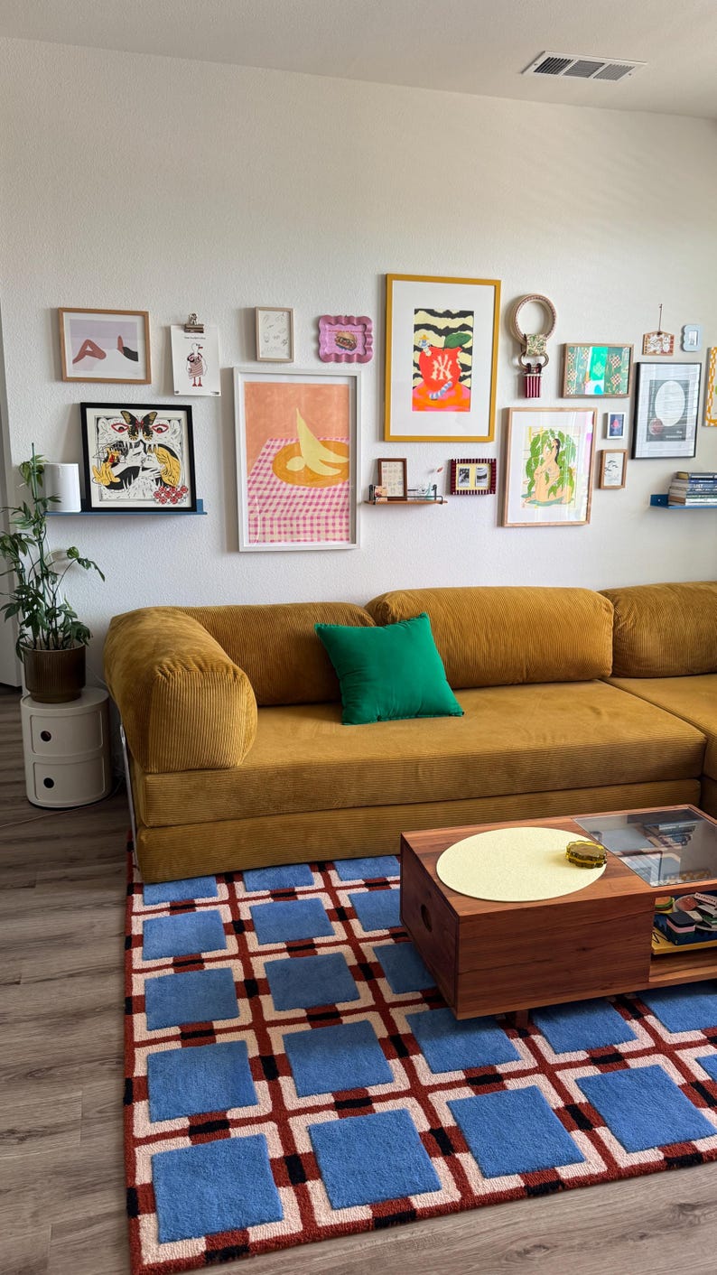 Op de afbeelding: Een mosterdgeel corduroy hoekbank met een groen kussen, een houten salontafel en een blauw en rood patroon tapijt. Verschillende ingelijste kunstwerken hangen aan de witte muur boven de bank.