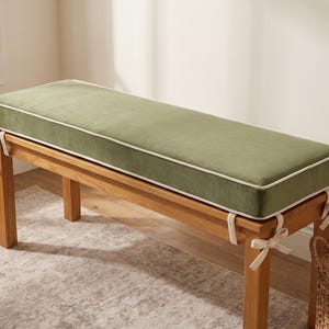 May include: A rectangular bench cushion in a muted sage green velvet fabric with cream-coloured piping along the edges. The cushion is secured to a wooden bench frame with matching cream-coloured fabric ties at each of the four corners.