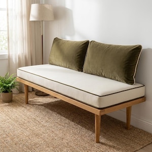 May include: A wooden bench featuring a long, off-white seat cushion with dark green piping. Two rectangular, olive green velvet pillows are positioned against the back. The bench is supported by four tapered wooden legs and has a clean, simple frame.