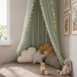 May include: A child's reading corner featuring a sage green canopy with fairy lights. It has cloud, star, and flower-shaped cushions, wooden blocks, and a stuffed rabbit. The bed is a round, corduroy-covered cushion.