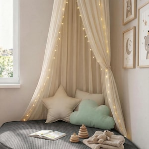 May include: A child's reading corner featuring a cream canopy with fairy lights. A grey, corduroy-textured floor cushion is furnished with star and cloud pillows, a book, wooden toys, and a teddy bear. Framed artwork decorates the wall.