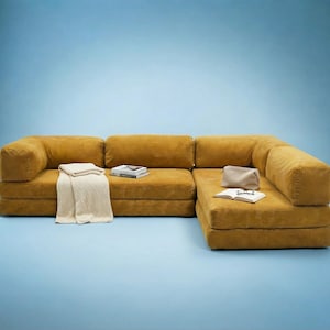 Op de afbeelding: Mosterdgele modulaire bank met een ribfluwelen textuur. De bank is in een L-vorm geplaatst en bevat een cr&egrave;mekleurige deken, een boek en een kleine tas. De achtergrond is effen lichtblauw.
