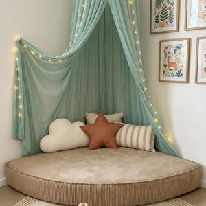 May include: A children's reading nook featuring a round, corduroy floor cushion, a mint green canopy with fairy lights, and decorative pillows. A woven rug, wooden toys, and framed art create a welcoming space.