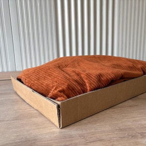 Op de afbeelding: Een gevouwen, roestkleurig textiel, mogelijk een deken of kledingstuk, ligt in een eenvoudige kartonnen bak. De stof heeft een geribbelde textuur. De bak staat op een houten oppervlak, met een witte, gegroefde muur op de achtergrond.