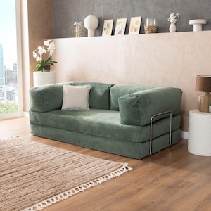 May include: A green corduroy sofa with a square shape and a light-colored square pillow. The sofa has a metal frame and is placed on a wooden floor next to a brown rug. The room has a neutral color palette.