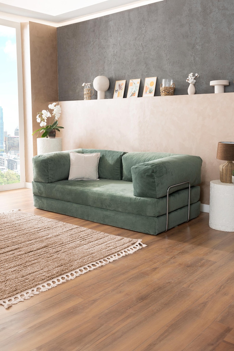 May include: A green corduroy sofa with a square shape and a light-colored square pillow. The sofa has a metal frame and is placed on a wooden floor next to a brown rug. The room has a neutral color palette.