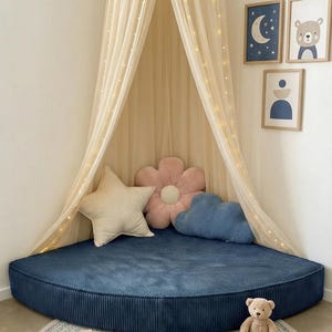 May include: A children's reading nook featuring a navy blue corduroy floor cushion, a cream canopy with fairy lights, and decorative pillows. A round rug, a teddy bear, and framed art complete the space. The room is decorated in soft, neutral tones.