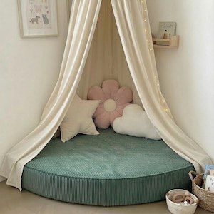 May include: A children's reading corner featuring a green corduroy floor cushion, a cream canopy, and decorative pillows shaped like a star, flower, and cloud. A framed animal print and fairy lights create a cosy space.