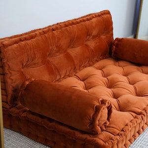 May include: A burnt orange velvet floor sofa featuring a tufted design with deep button detailing. The piece includes a rectangular backrest and a flat seat cushion, accompanied by two cylindrical bolster pillows placed on each side of the seat.