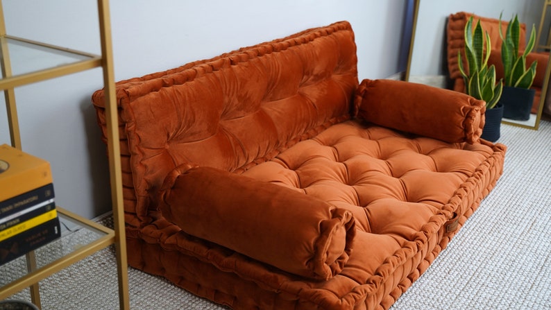 May include: A burnt orange velvet floor sofa featuring a tufted design with deep button detailing. The piece includes a rectangular backrest and a flat seat cushion, accompanied by two cylindrical bolster pillows placed on each side of the seat.