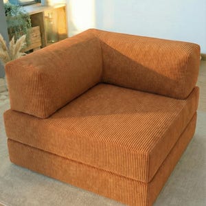 May include: A modular orange corduroy sofa. The sofa has a low profile and a simple, modern aesthetic. The corduroy fabric has a ribbed texture. The sofa is placed on a neutral-coloured rug. The sofa is suitable for a small living space.