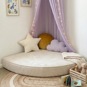 Può includere: Un'accogliente area giochi per bambini con un cuscino rotondo da pavimento in velluto a coste color crema. Una tenda da letto lavanda con luci di fata è appesa sopra. Cuscini decorativi a forma di stella, nuvola e rotondi nei colori crema, lavanda e oro. Una cassa di legno contiene giocattoli e libri.