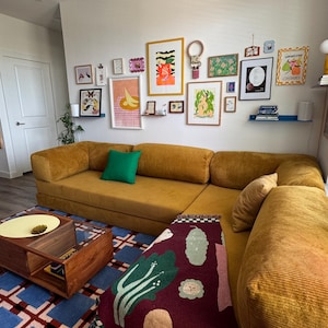 Op de afbeelding: Een mosterdgele hoekbank met een groen kussen en een patroon plaid. Een blauw en rood geometrisch tapijt en een houten salontafel staan voor de bank. Diverse ingelijste kunstwerken sieren de witte muur.