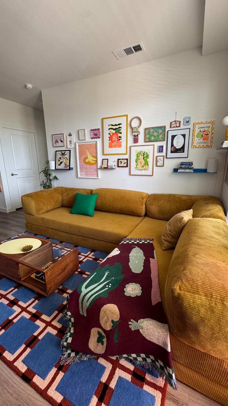 Op de afbeelding: Een mosterdgele hoekbank met een groen kussen en een patroon plaid. Een blauw en rood geometrisch tapijt en een houten salontafel staan voor de bank. Diverse ingelijste kunstwerken sieren de witte muur.
